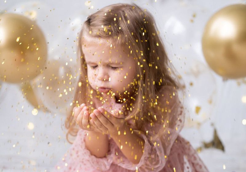 Una niña con los ojos cerrados soplando serpentina dorada.